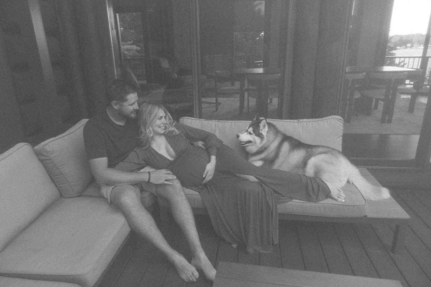 A relaxed couple lounges on an outdoor sofa with their husky dog in a casual black and white photo.
