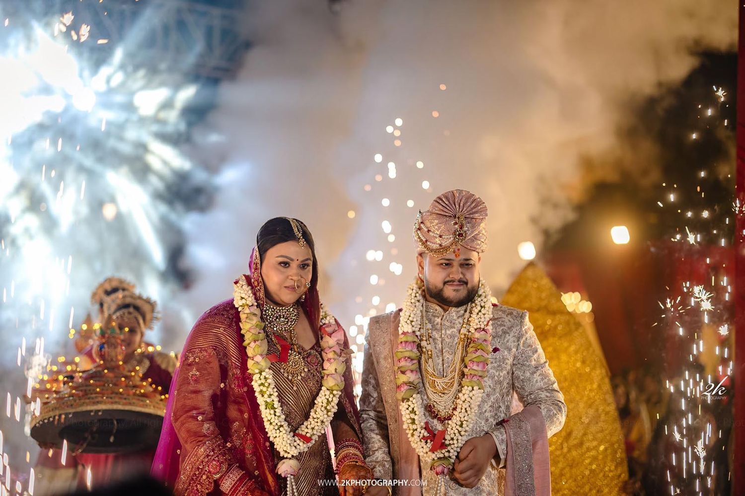 Indian couple celebrates their wedding ceremony with fireworks, garlands and traditional attire against nighttime backdrop.