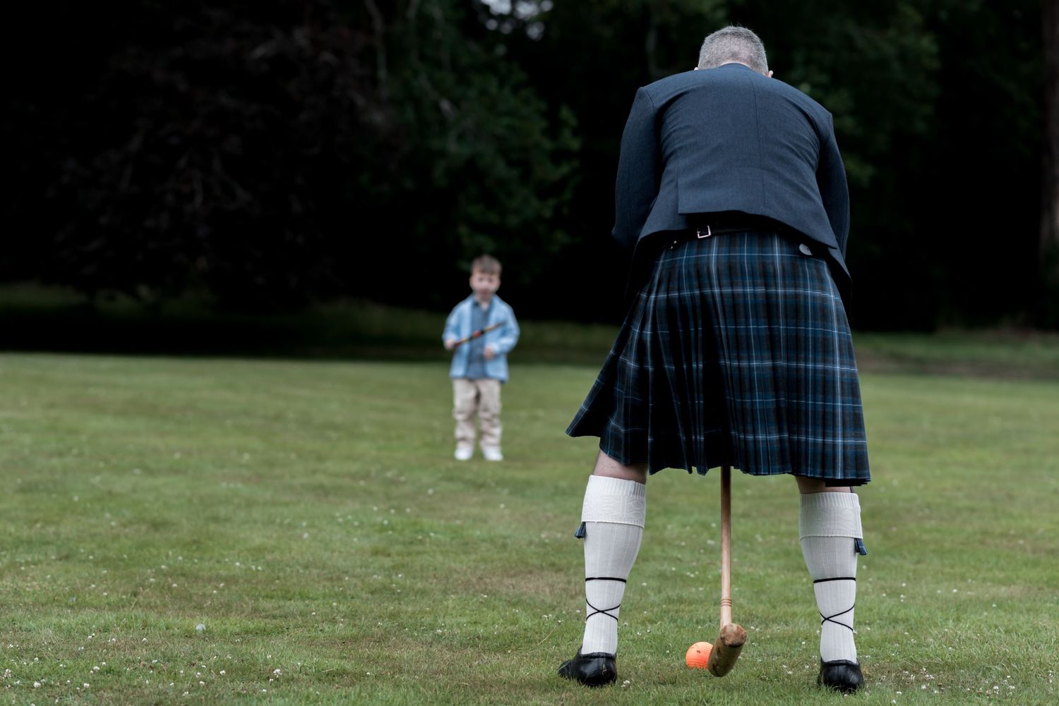 A person wearing traditional Scottish kilt and socks stands in a grassy field facing someone small in the distance.