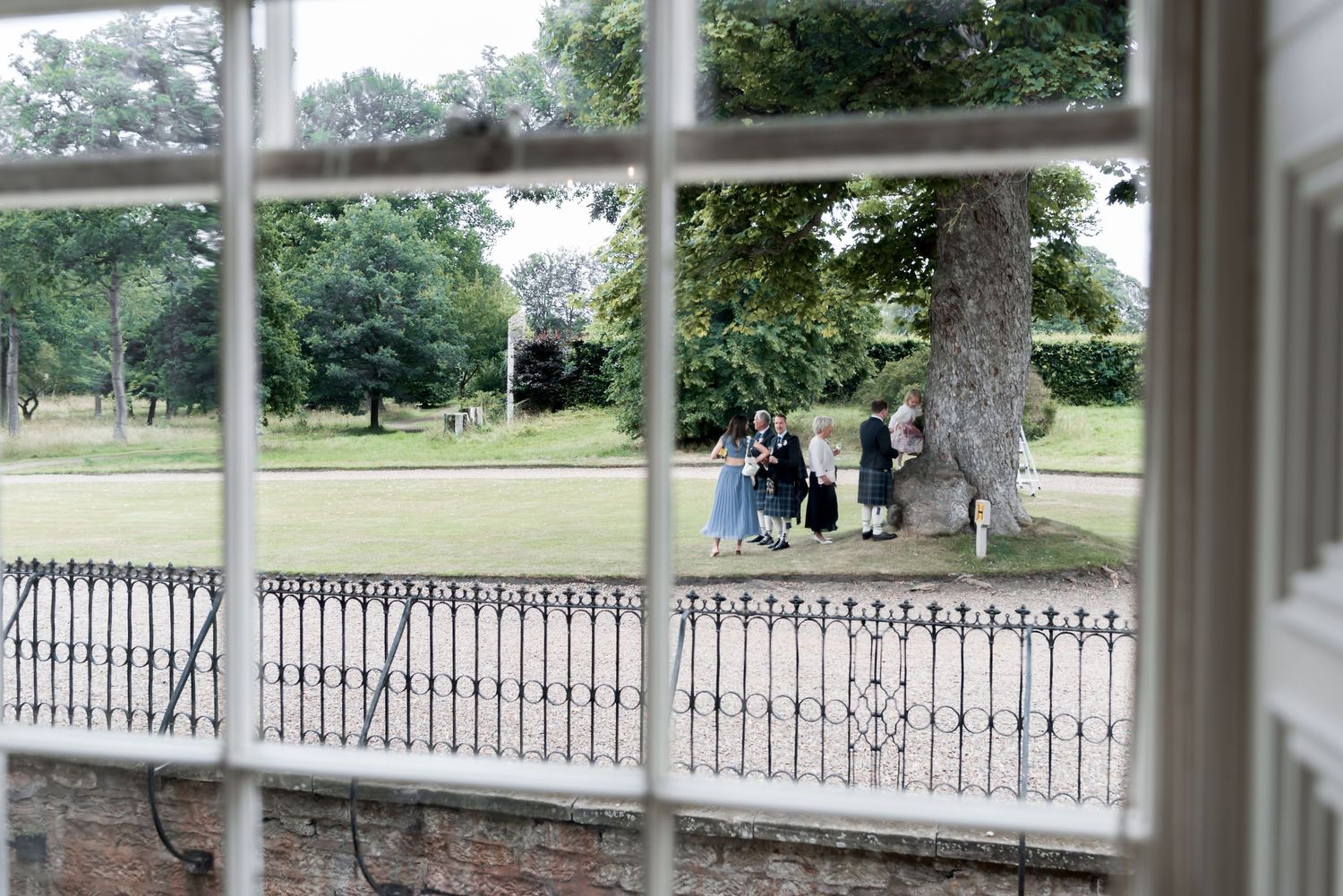 View through window panes of people sitting under tree by iron railing in peaceful park setting.