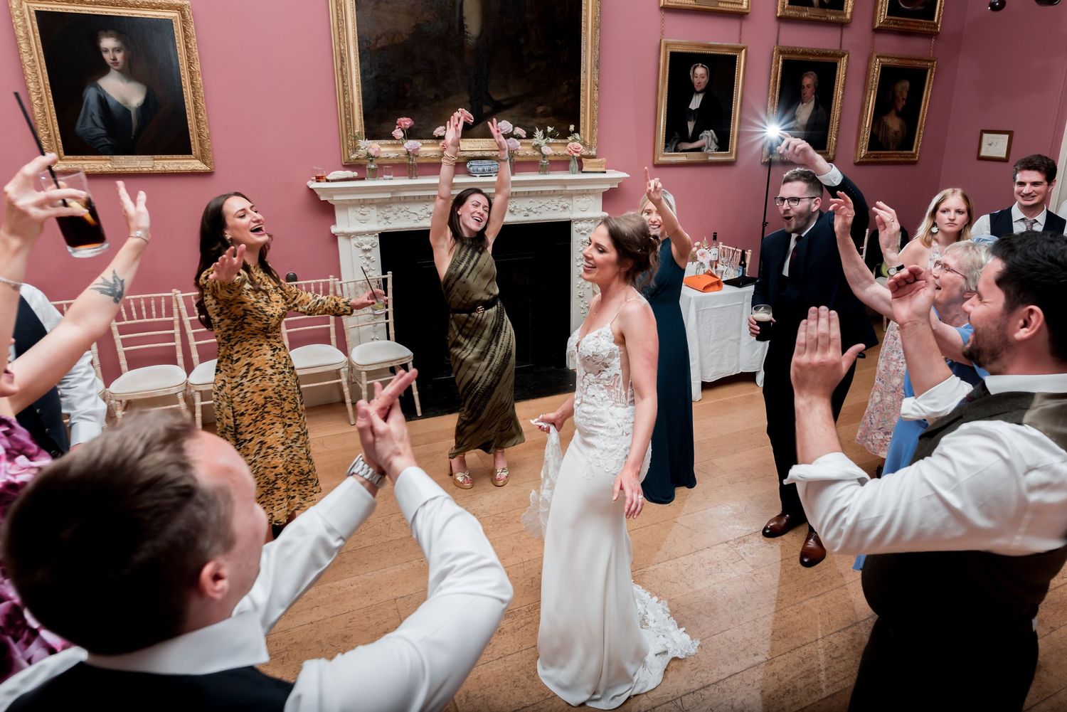 Wedding guests dancing energetically together in an elegant gallery room with pink walls and ornate framed artwork.