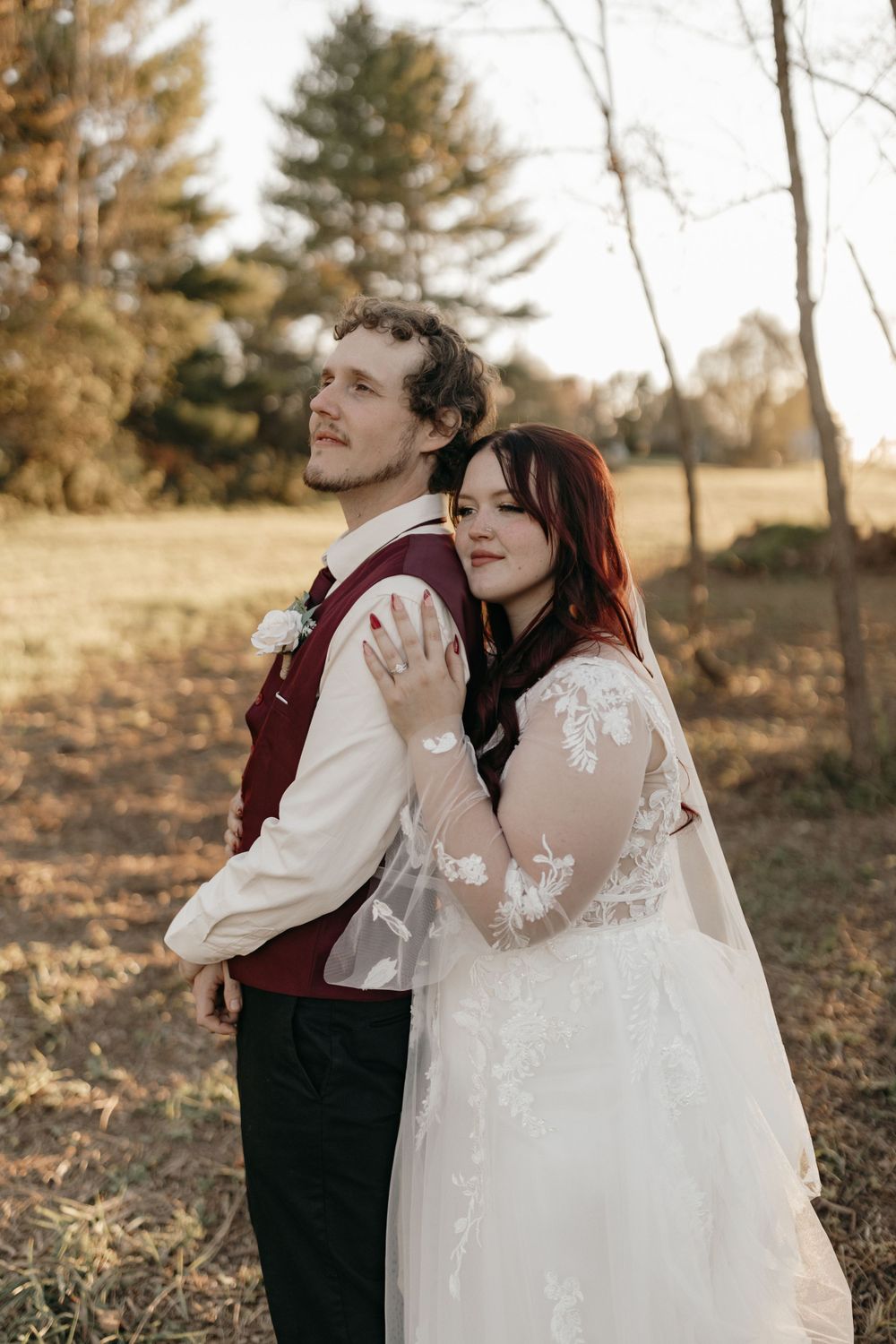 Newlyweds embrace in autumn sunlight wearing burgundy vest and white lace gown.