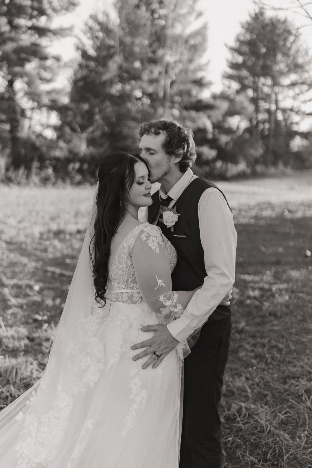 Black and white portrait of romantic wedding moment with pine trees in background.