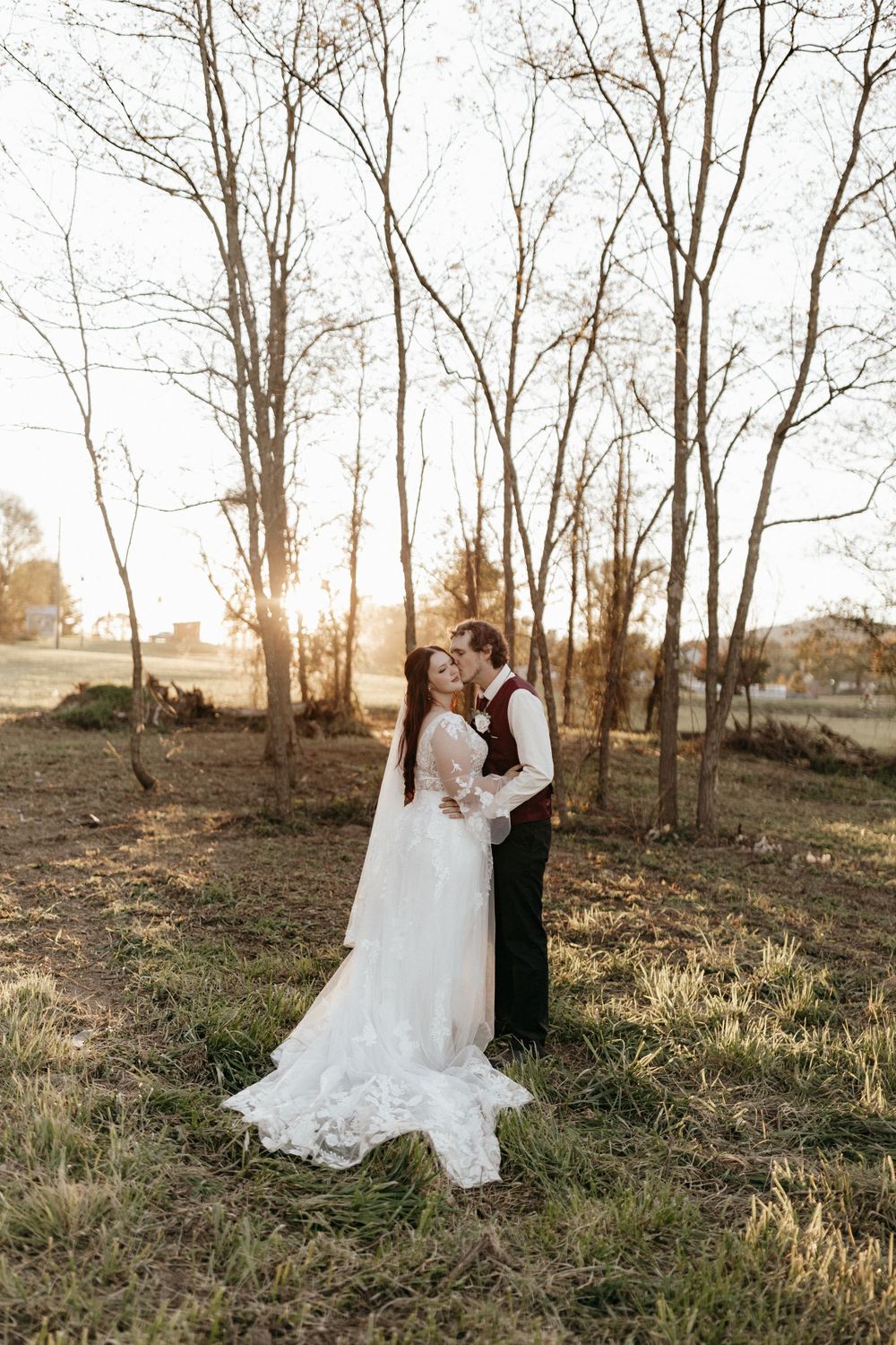 A romantic sunset wedding photo of newlyweds embracing among bare trees in a rustic outdoor setting.