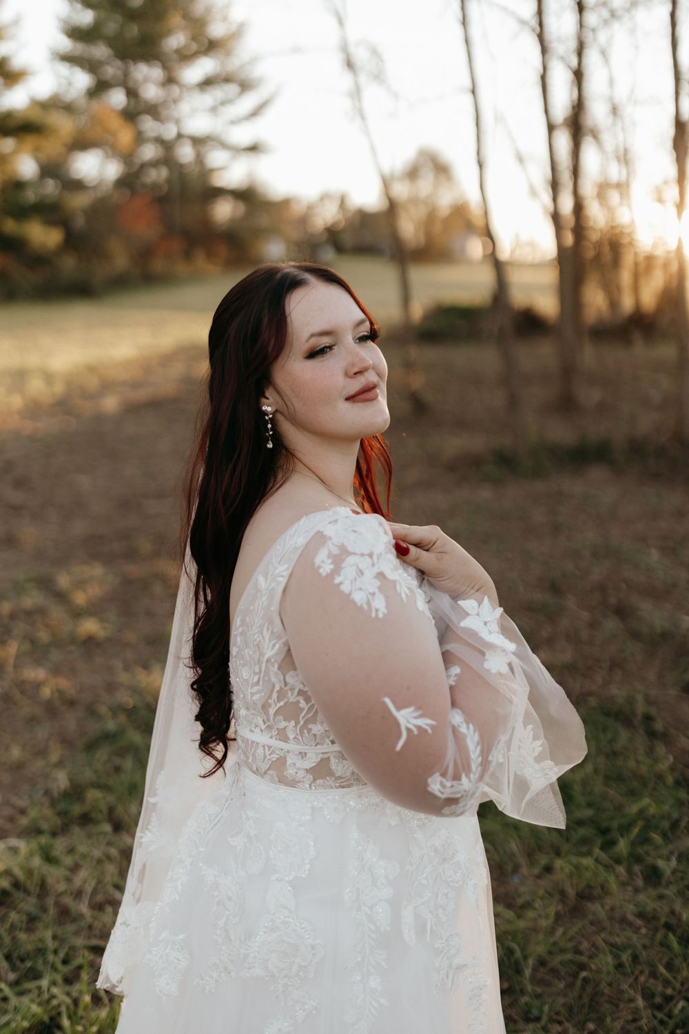 A bride in a lace wedding gown poses in an outdoor autumn setting at golden hour.