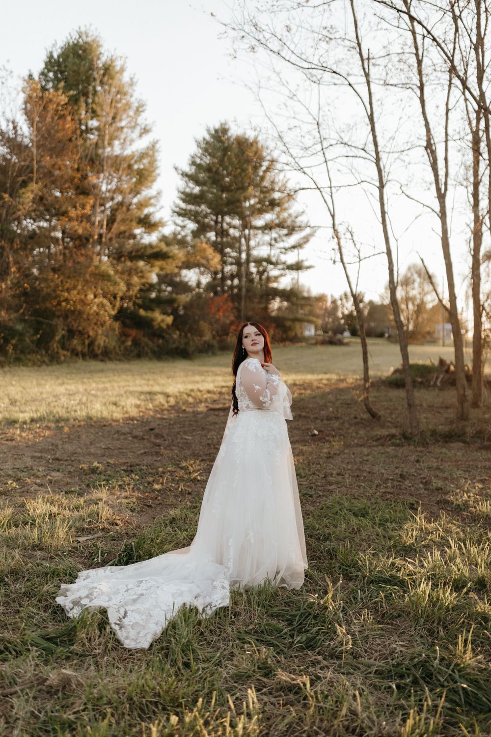 A bride in a flowing white lace dress stands in a woodland setting during sunset.