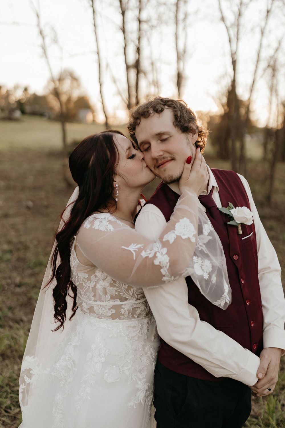 A tender moment between newlyweds sharing a kiss in front of bare trees at sunset.