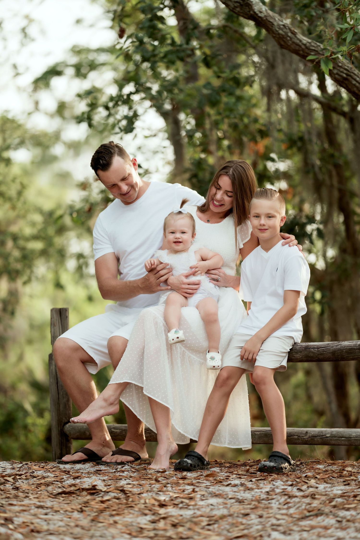 Family dressed in white sits together on a rustic wooden fence in a wooded outdoor setting during a photo session.