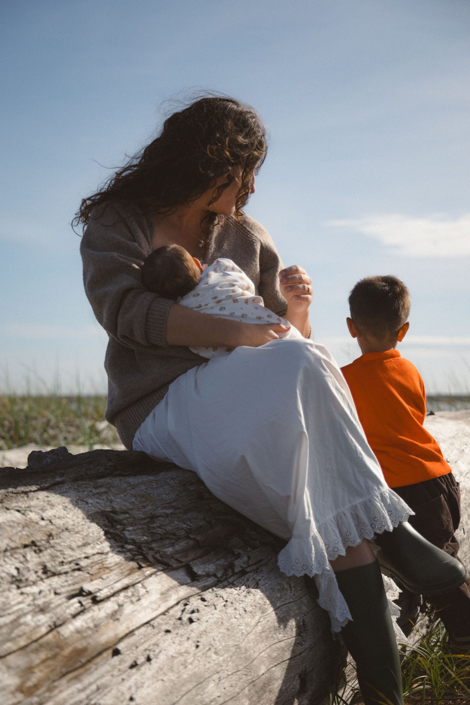 Mom holding her newborn baby while sitting on a log at the beach and looking out to the ocean where her toddler is showing her something.