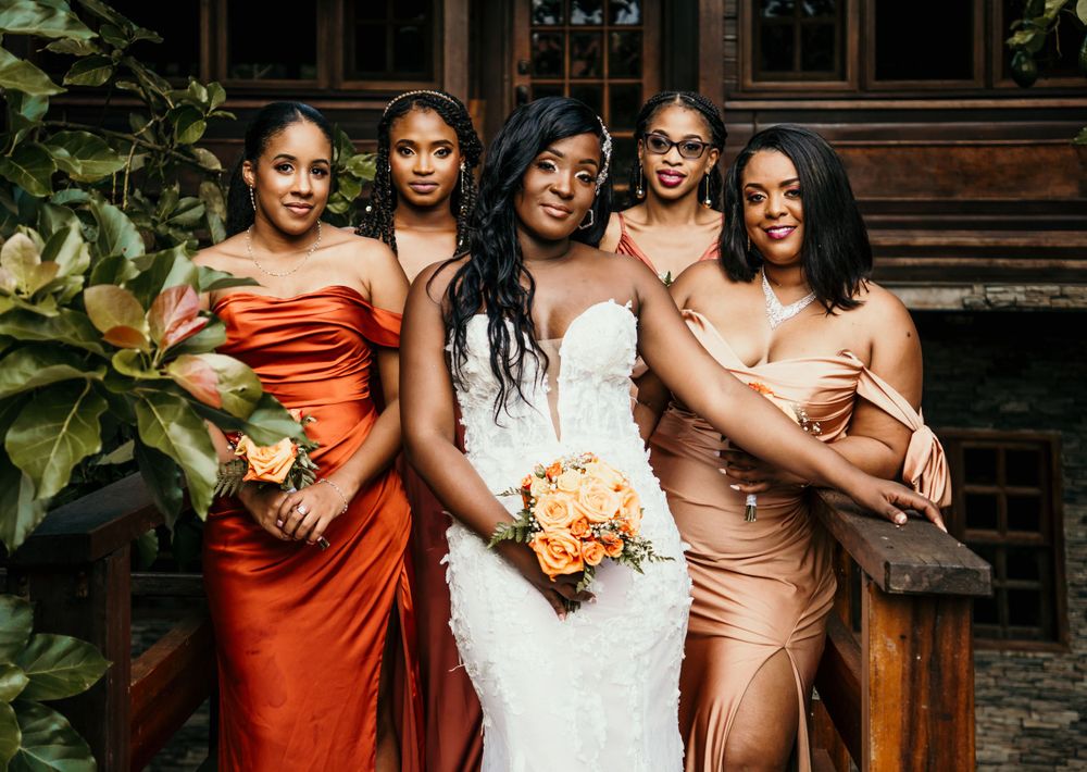 A bridal party in elegant orange and pink dresses poses together outdoors.