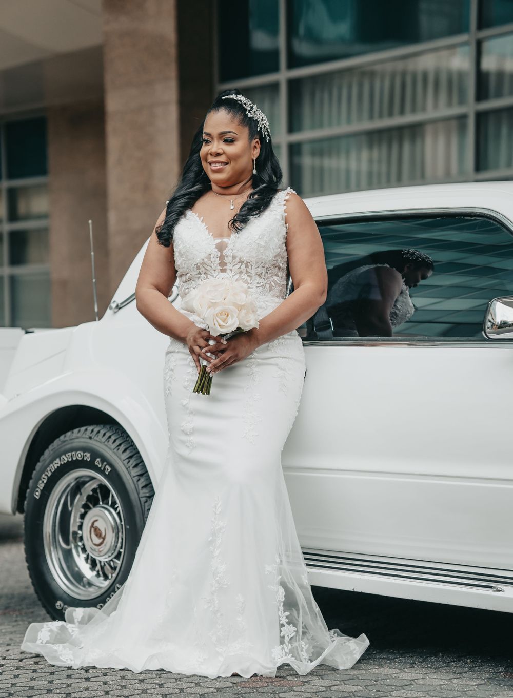 A bride in a fitted lace wedding gown poses beside a white limousine.