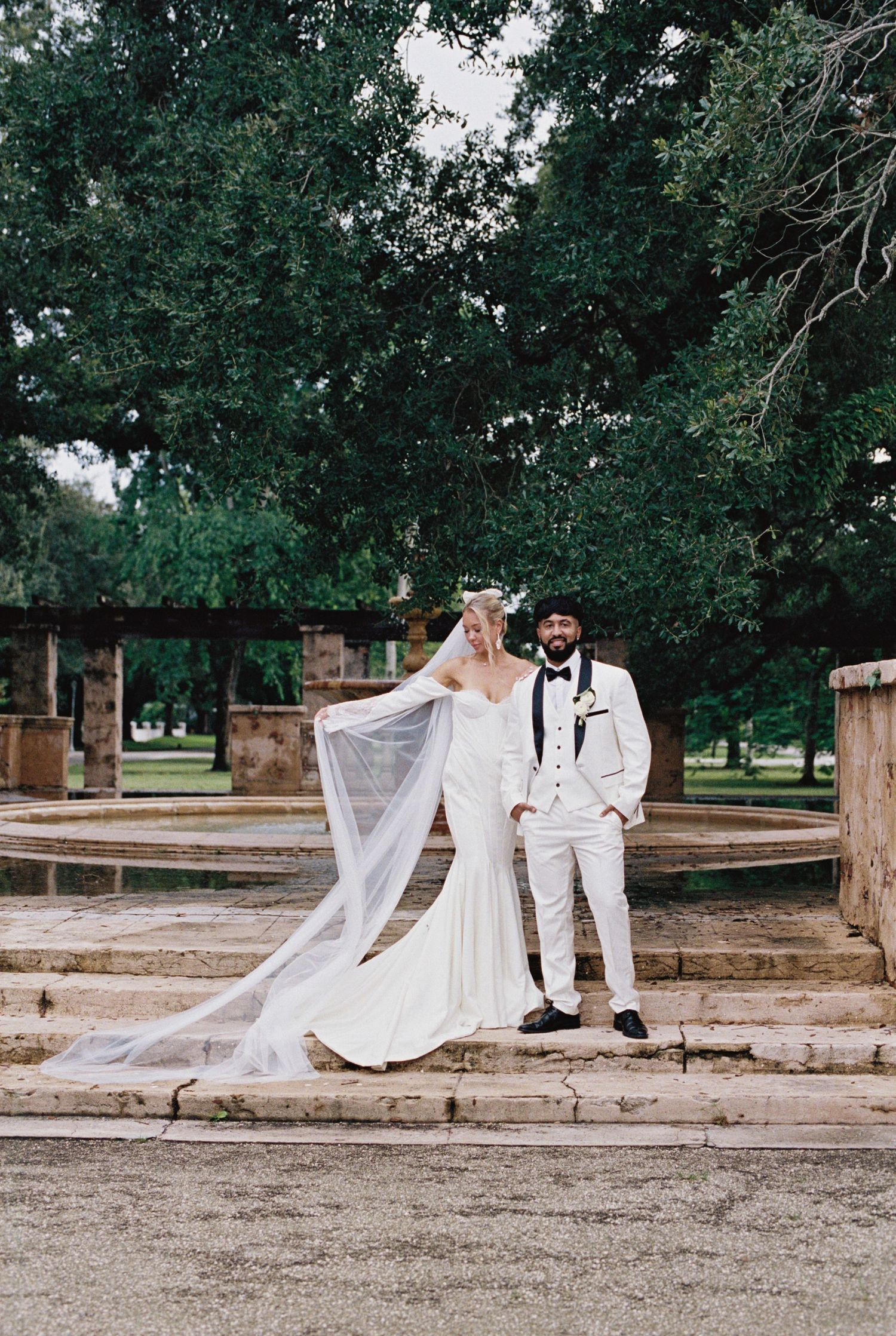 A wedding couple stands on stone steps surrounded by lush green trees in an outdoor venue with a flowing bridal veil.