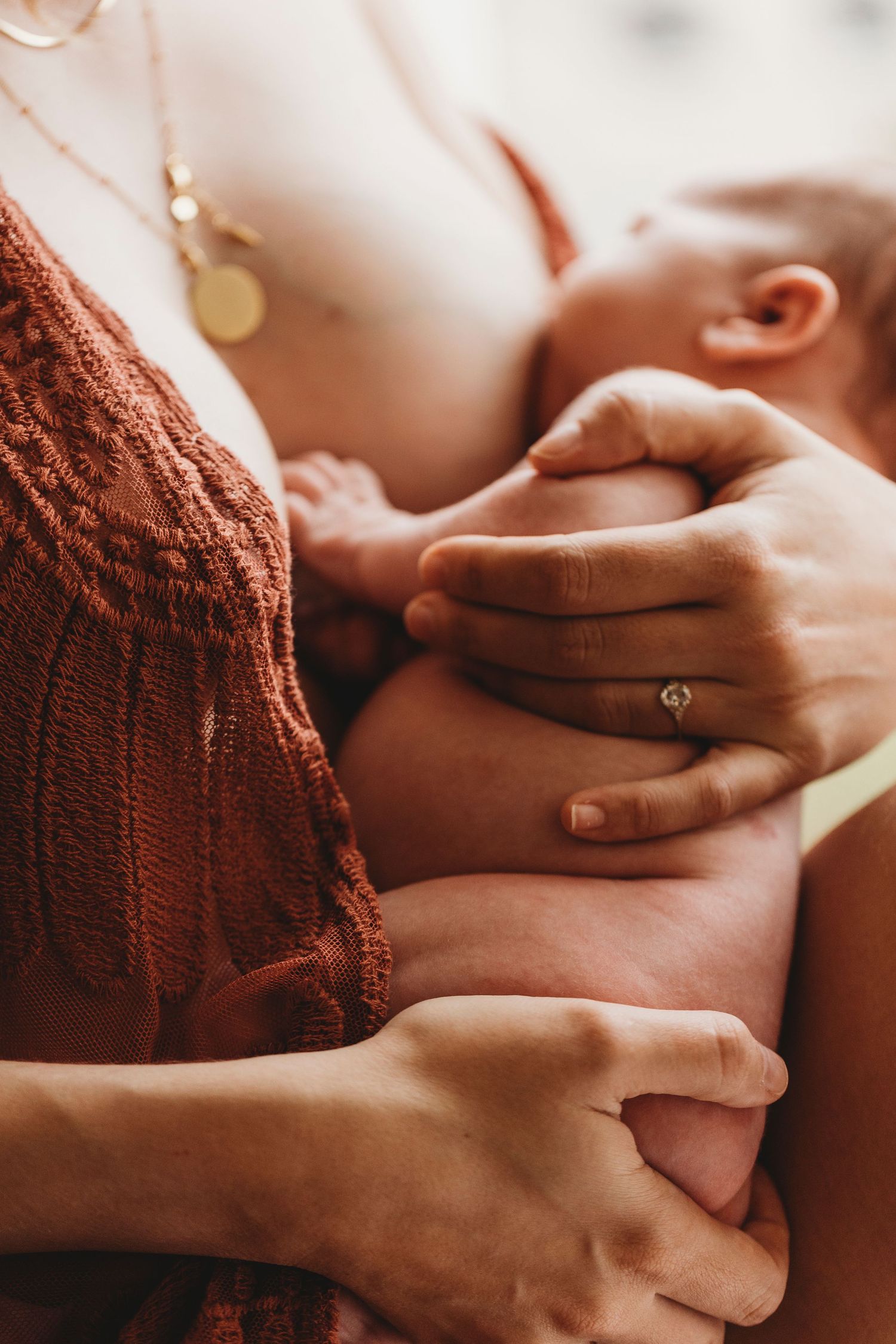 Closeup of hands tenderly holding an infant while breastfeeding, wearing a rust-colored knit sweater and delicate necklace.