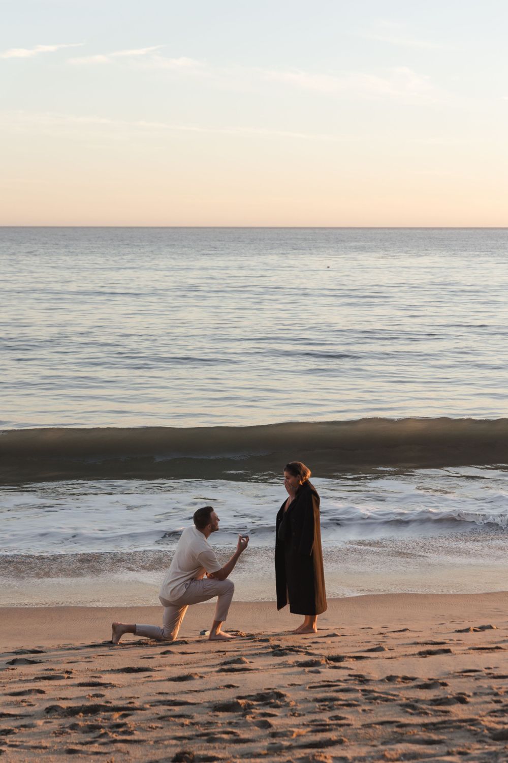 Two people share a romantic moment during sunset on a serene beach with calm ocean waves in the background.