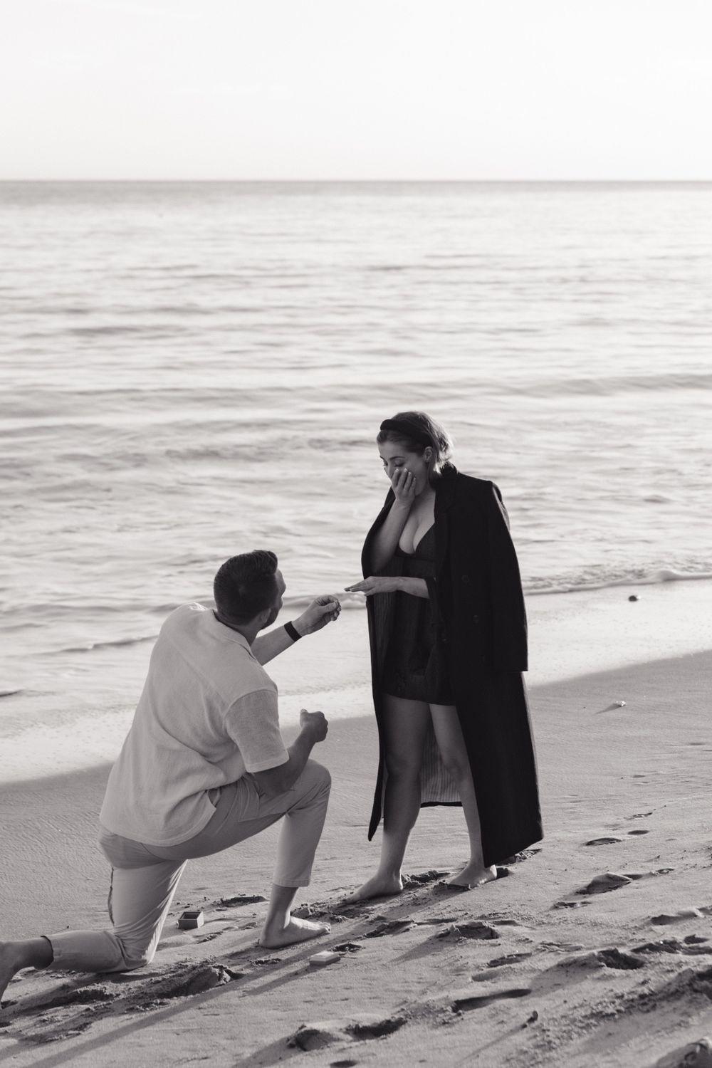 A romantic beach proposal at sunset with one person kneeling in the sand in black and white photography.