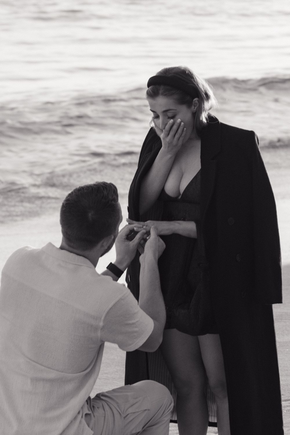 Black and white photo of a romantic beach proposal with person in white kneeling before someone in a dark outfit by the ocean.