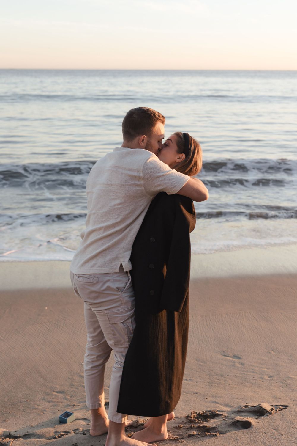 A couple shares an intimate moment on a sandy beach at sunset with ocean waves in the background.