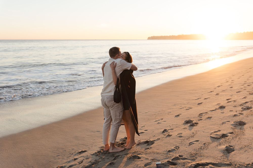 A couple embraces on a sandy beach at sunset with footprints in the sand and ocean waves in the background.