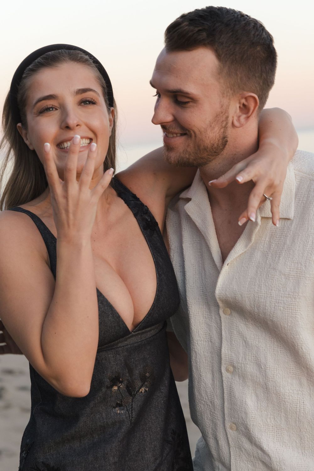 A couple celebrates their engagement with excited expressions at the beach.