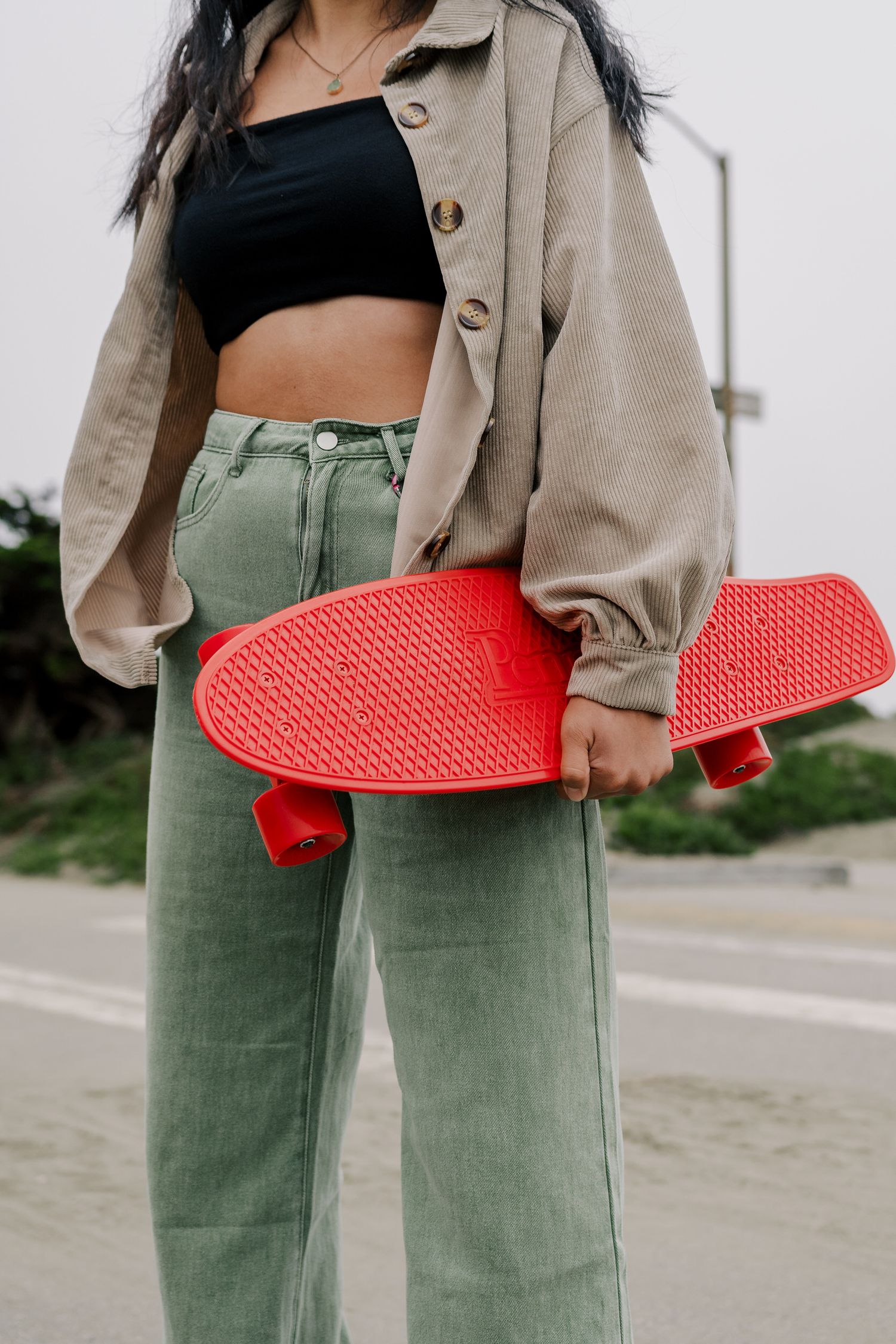 Someone in sage green pants and beige jacket holds a bright red skateboard deck against coastal backdrop.