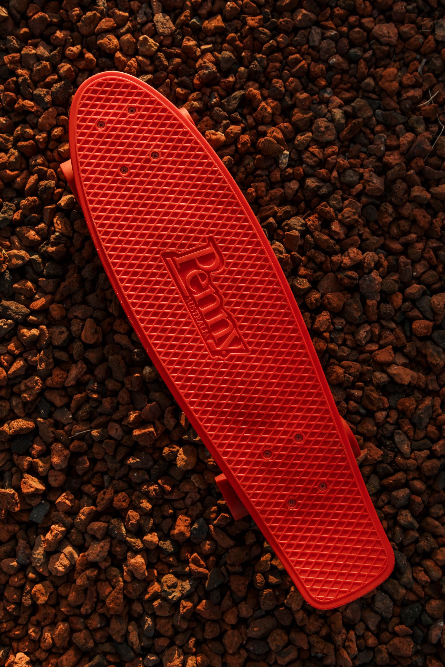 A bright red Penny skateboard deck rests on dark gravel stones creating striking visual contrast.