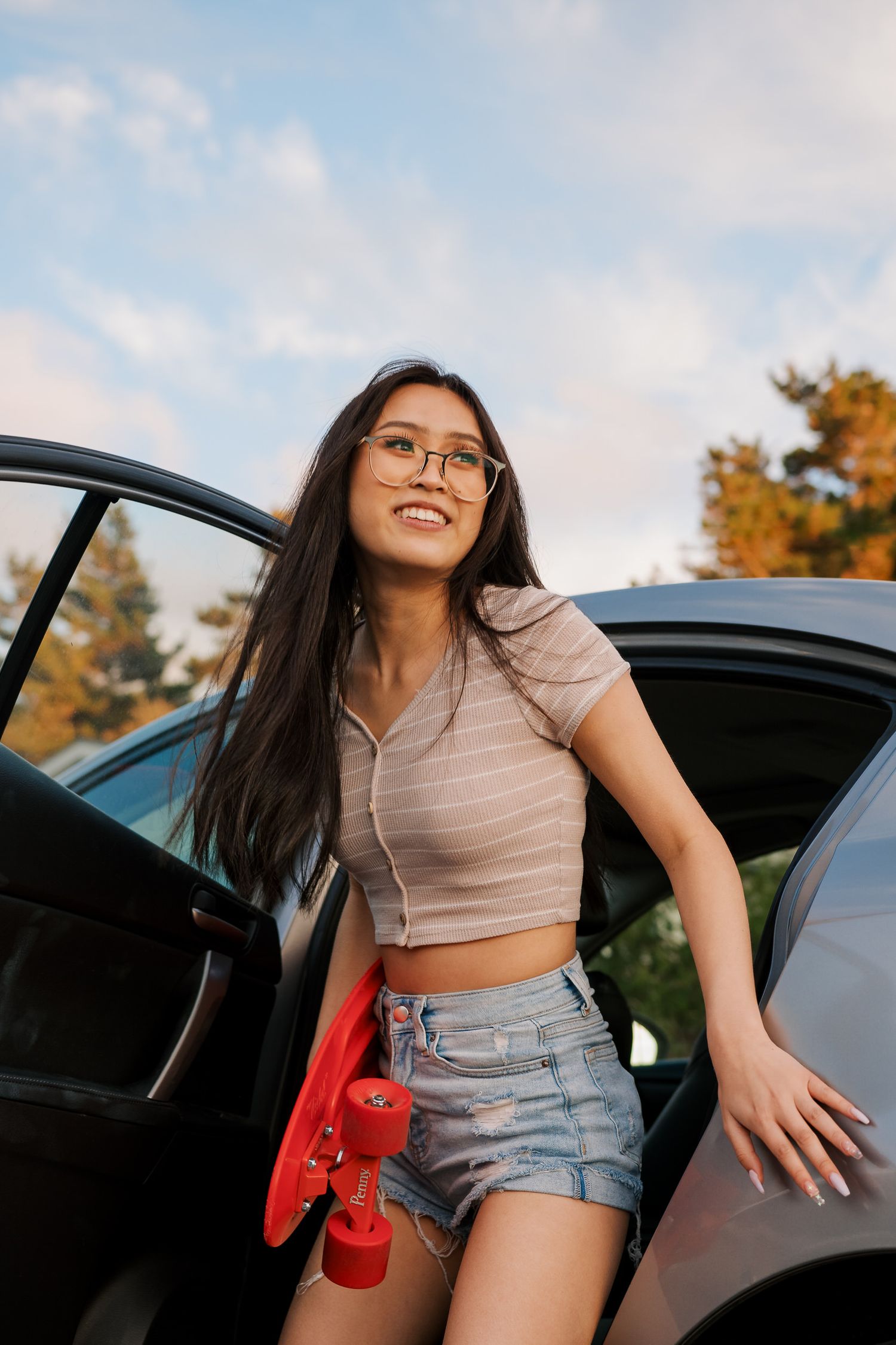 Someone getting out of a car wearing glasses, a beige crop top and denim shorts while holding a red skateboard.