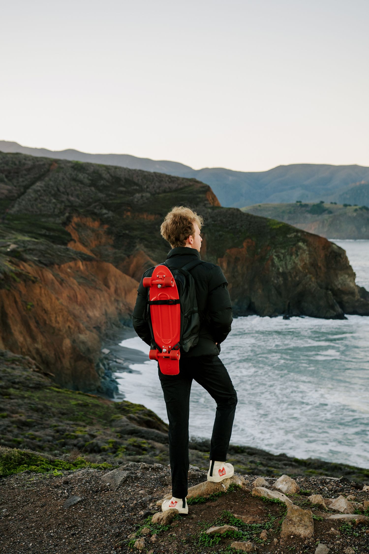 Person with red skateboard admires rugged coastal cliffs and crashing waves along a dramatic oceanside landscape at dusk.
