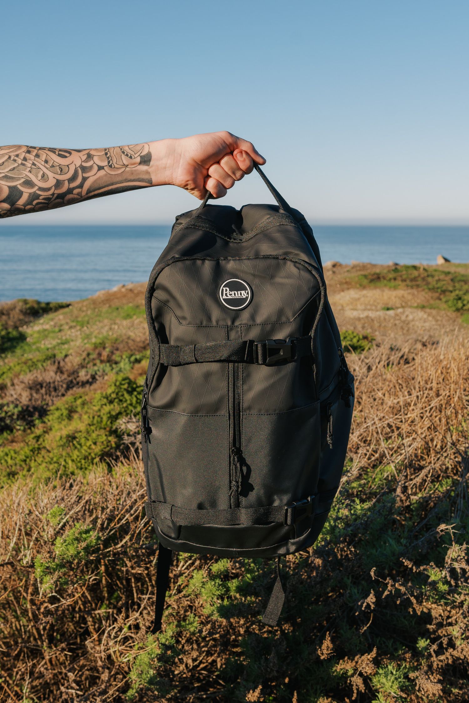 A tattooed arm holds up a black backpack with ocean and coastal cliffs visible in the background.