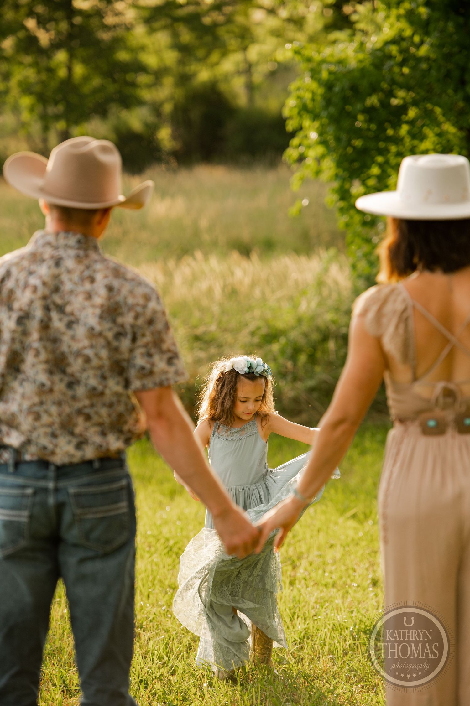 A young child in a blue dress dancing in a sunlit field while adults in cowboy hats watch.