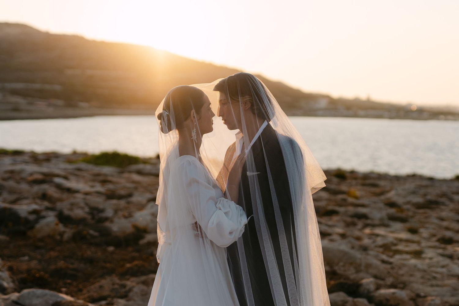 Two brides in white dresses embrace against a coastal sunset backdrop.