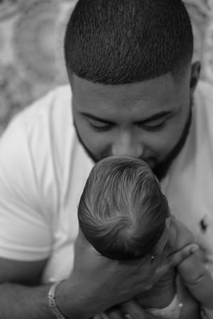 A tender black and white portrait of a parent gently cradling and kissing their newborn baby.