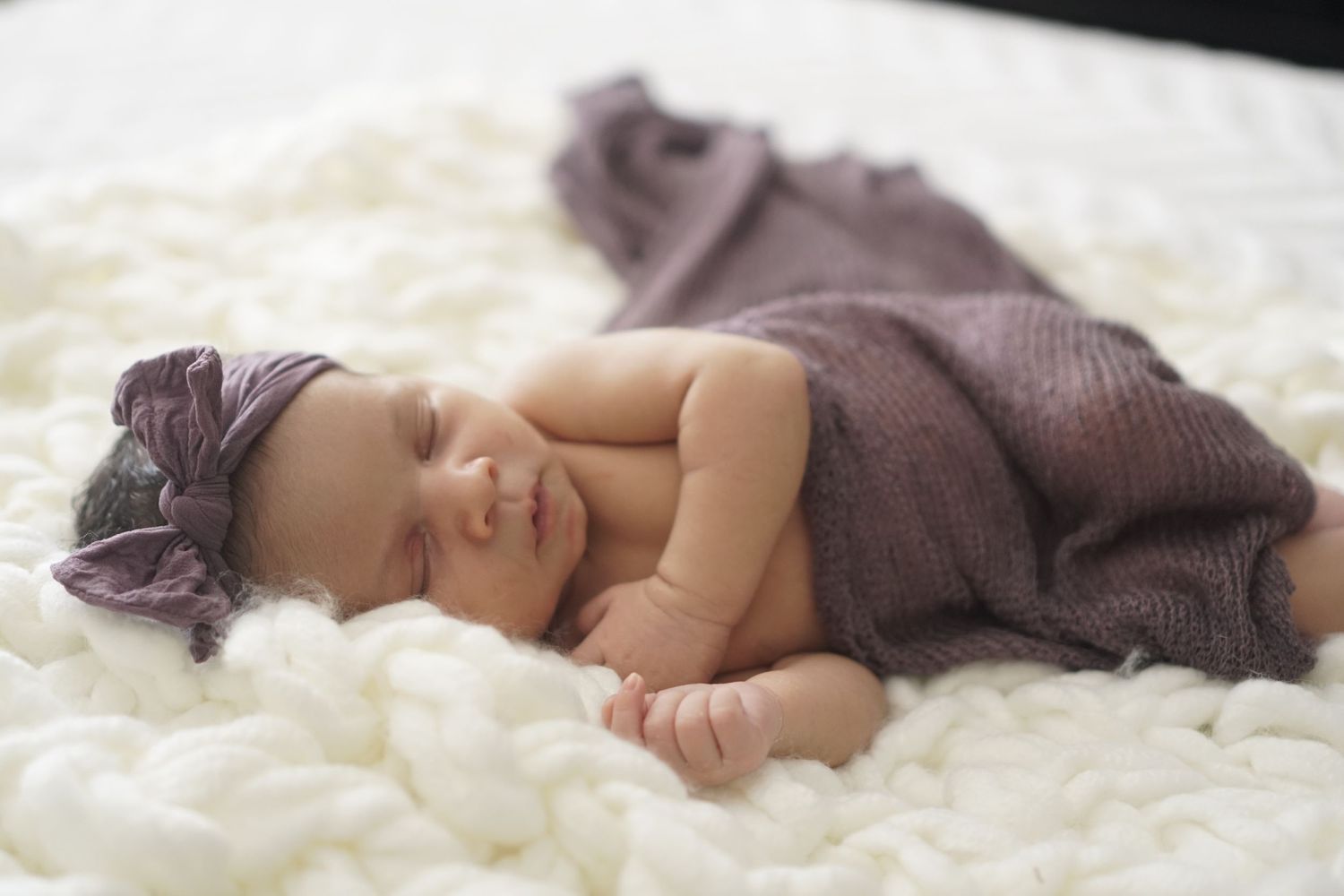 A sleeping newborn baby wearing a purple headband nestled in soft white blankets.