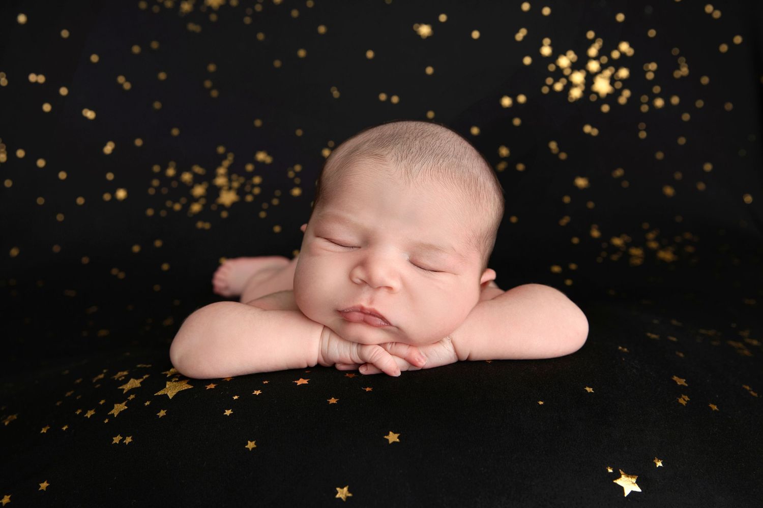 Newborn baby sleeping with crossed arms on black backdrop with gold sparkles.