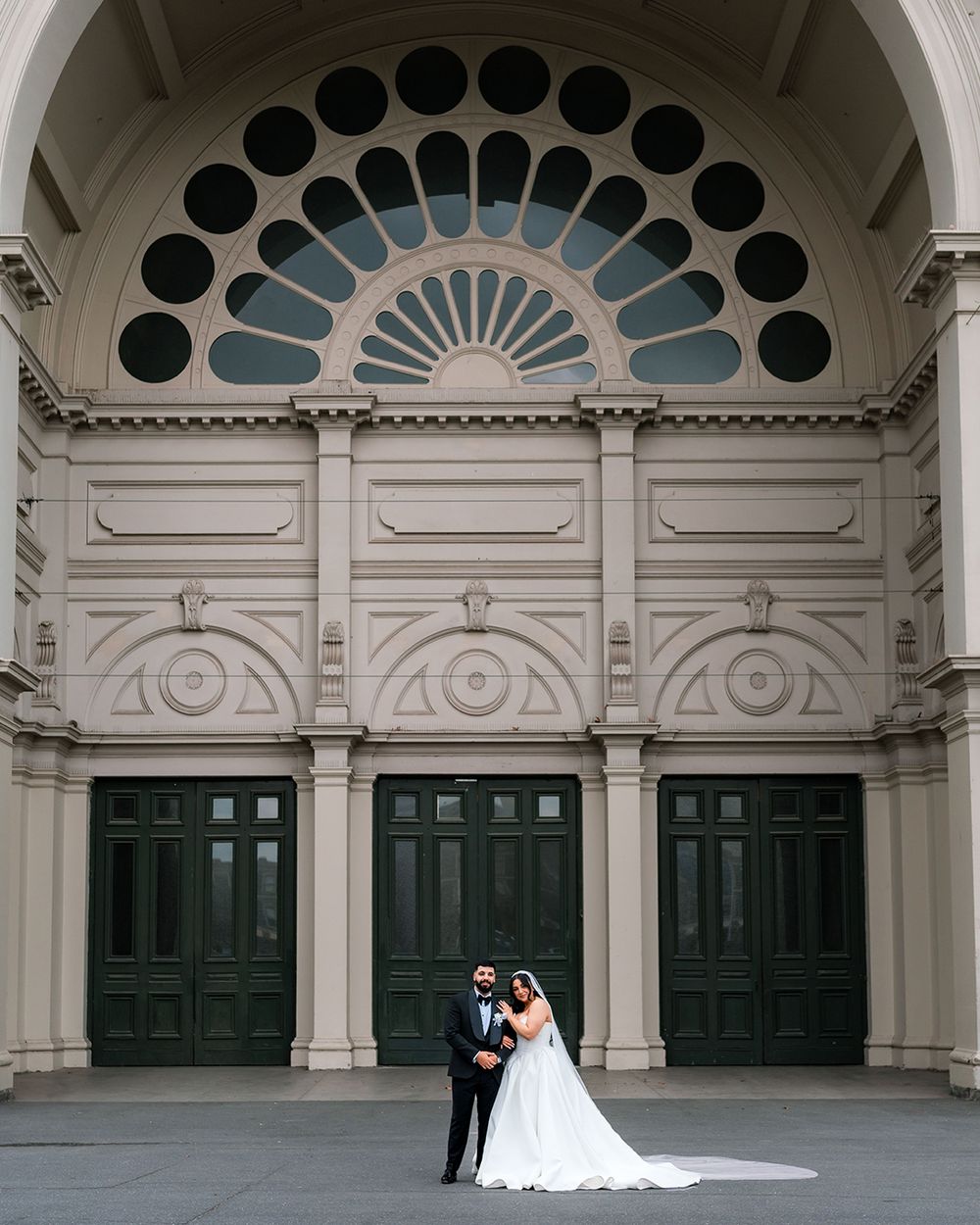 Couple in wedding attire stand in front of ornate white archways with green doors and decorative windows.
