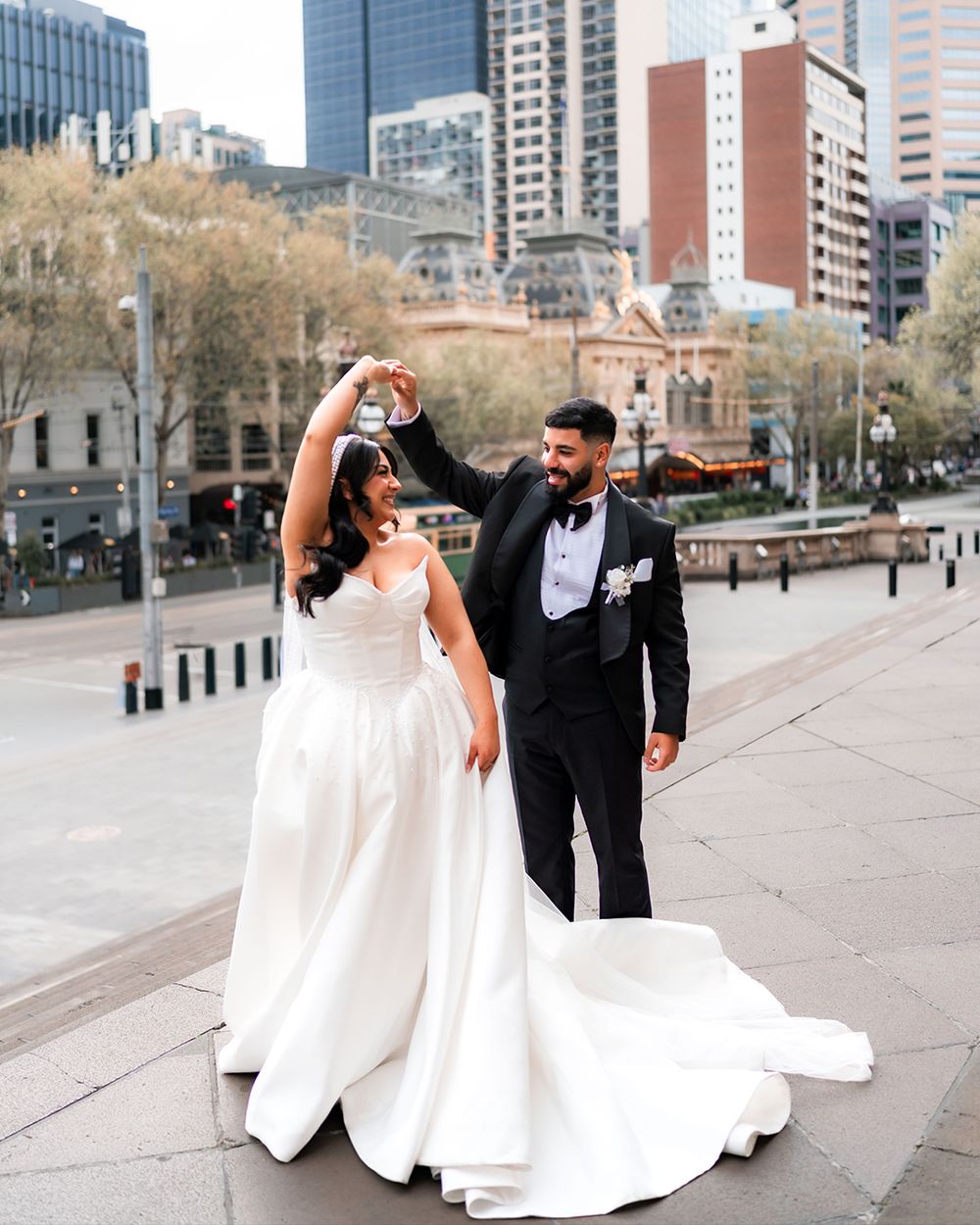 A couple in formal wedding attire dances and twirls on a city sidewalk with tall buildings in the background.
