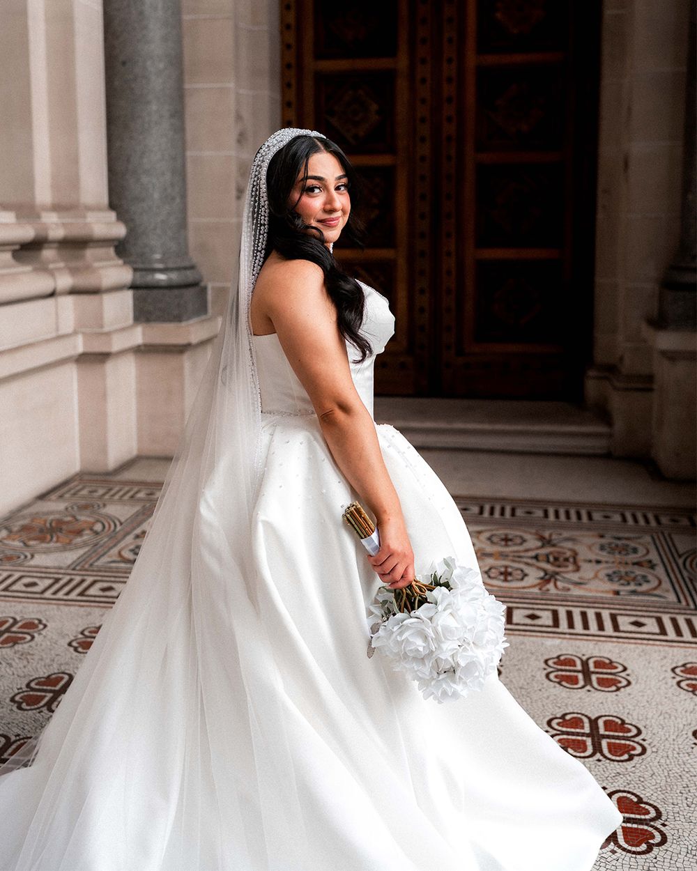 A bride in a flowing white wedding gown poses gracefully in an elegant hallway with ornate floor patterns.