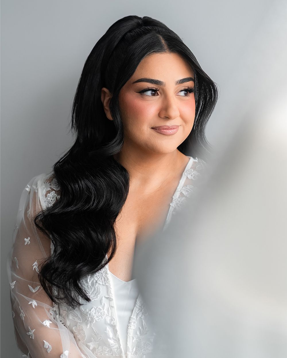 Portrait in soft lighting featuring long wavy black hair, white lace dress, and delicate sheer sleeve against grey backdrop.