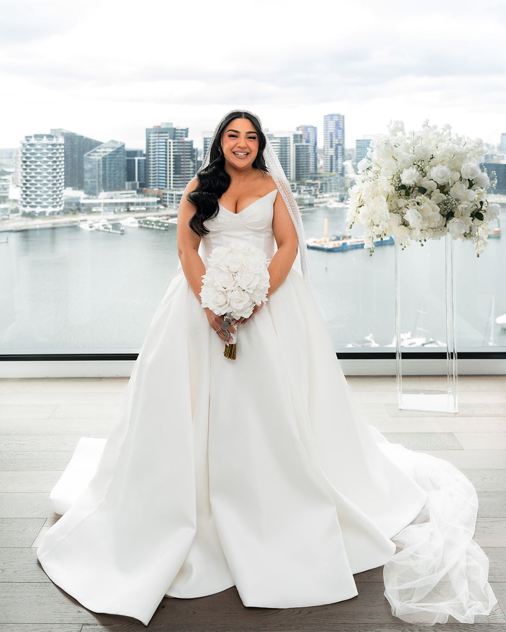 A bride in a flowing white wedding gown poses against a modern cityscape backdrop with white floral bouquet.