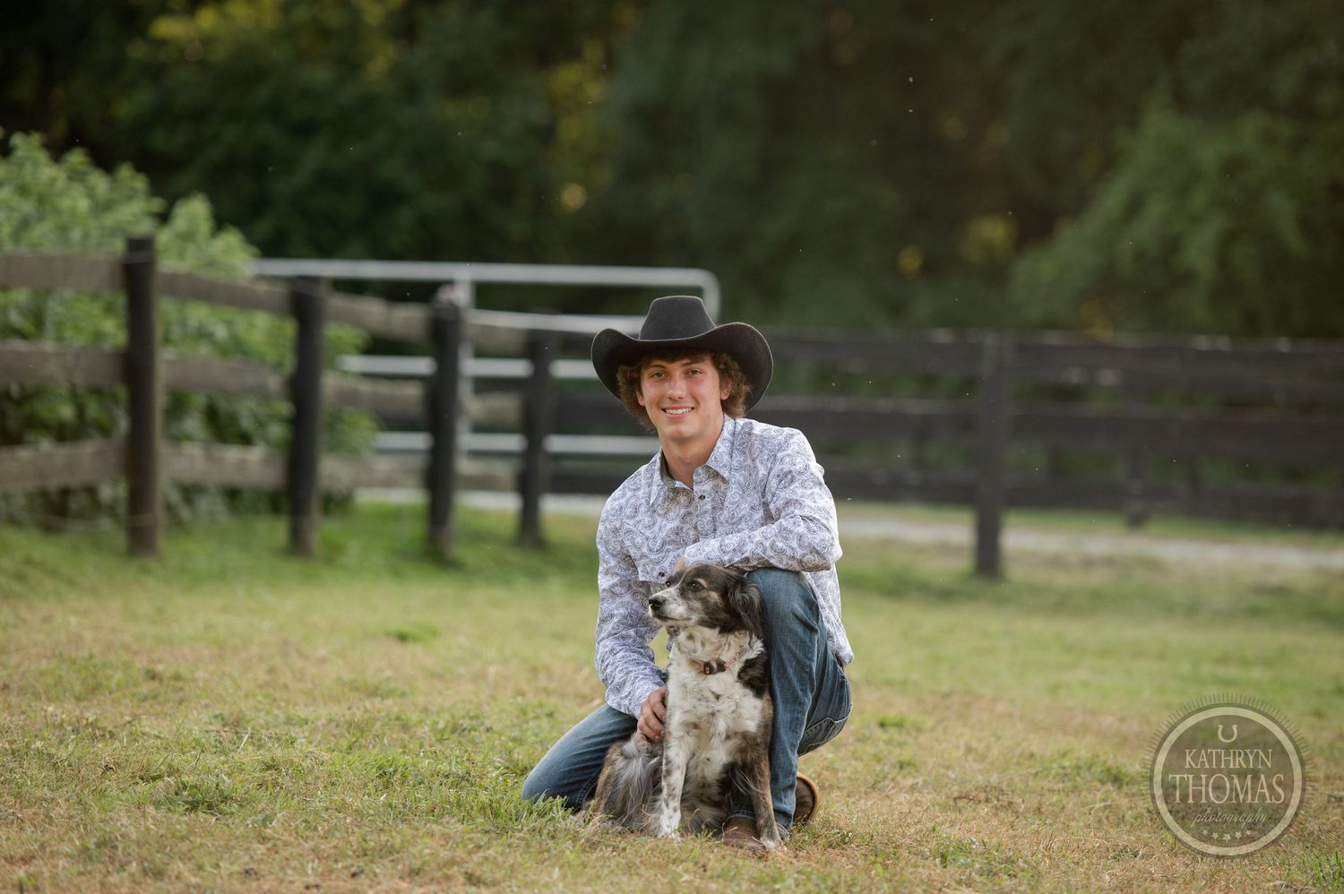 A person in western attire kneels beside their dog in a fenced pasture at sunset.