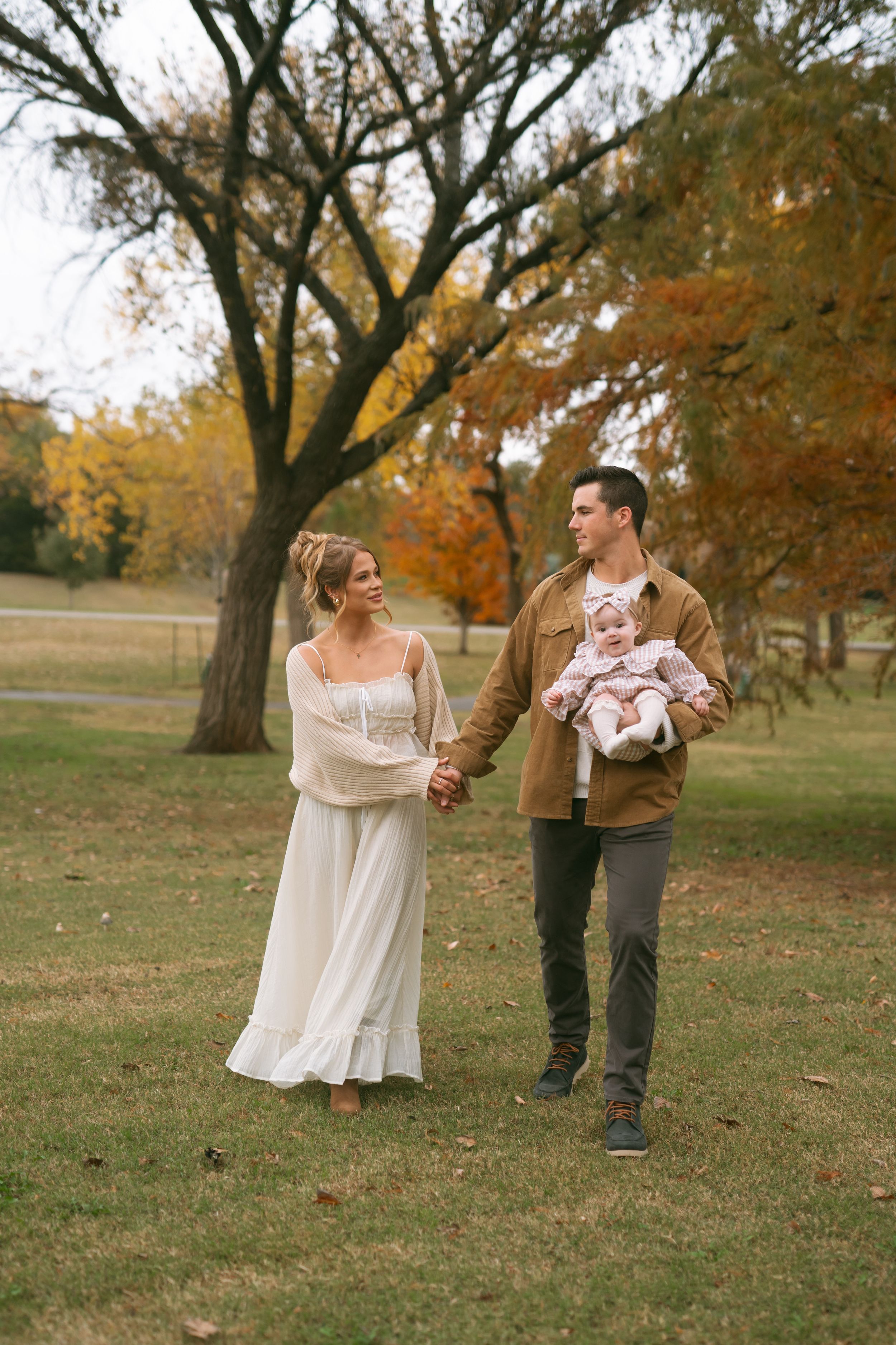 Family photoshoot in autumn park with parents and infant on grassy field with fall trees.
