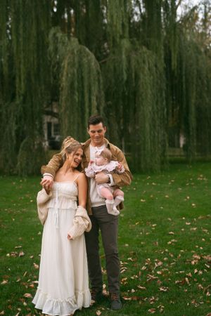 Family portrait session under weeping willow trees with parents and infant in casual attire.