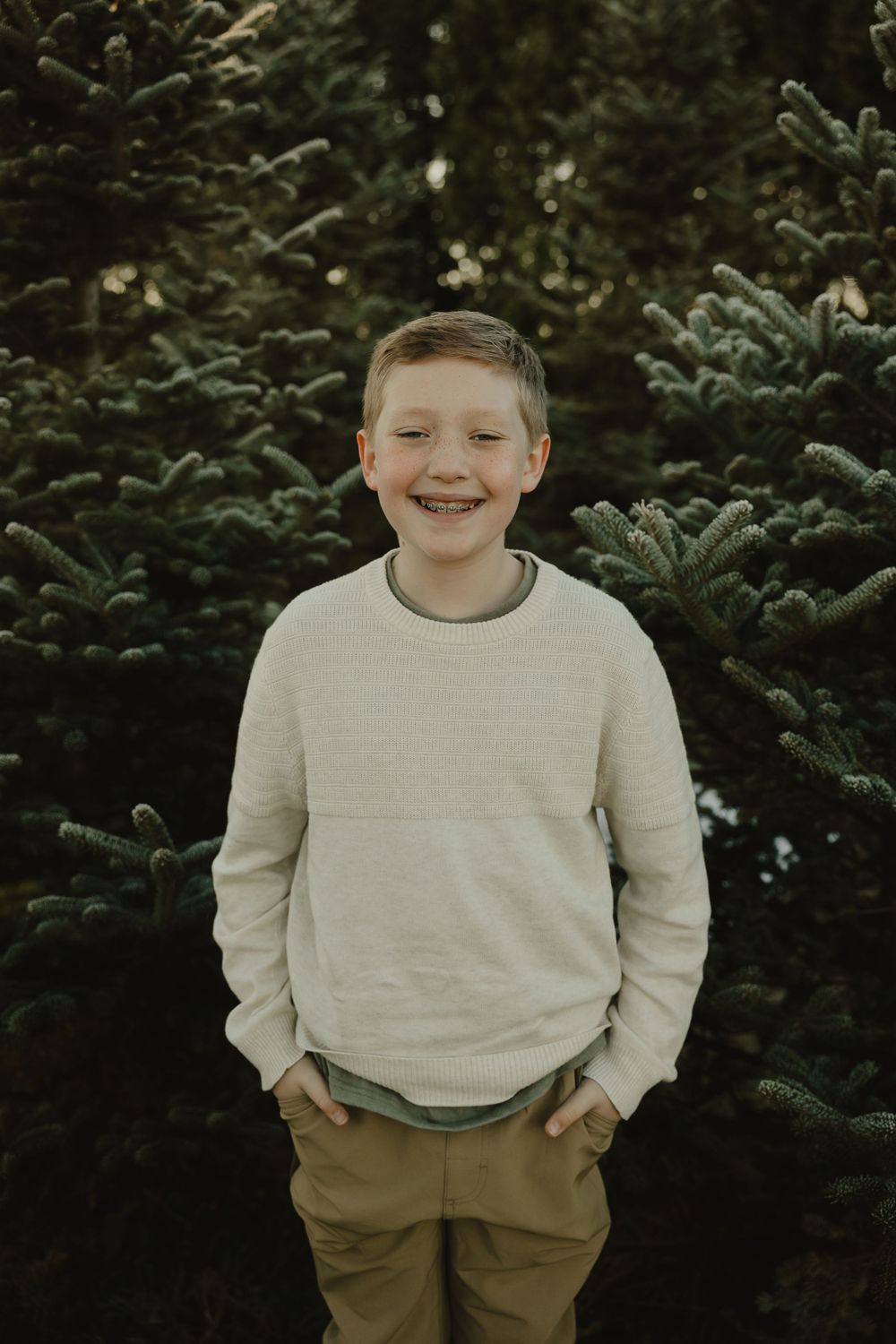 Young person in cream sweater smiling brightly against evergreen trees.