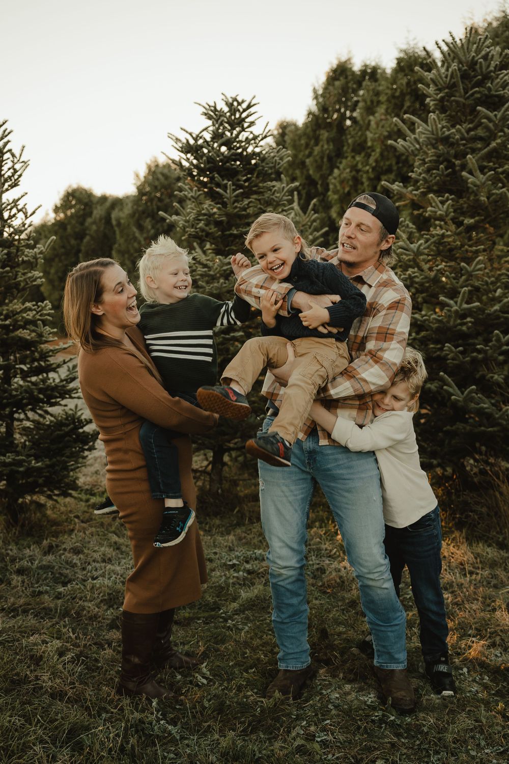 A family shares joyful moments together among evergreen trees at a Christmas tree farm during golden hour.