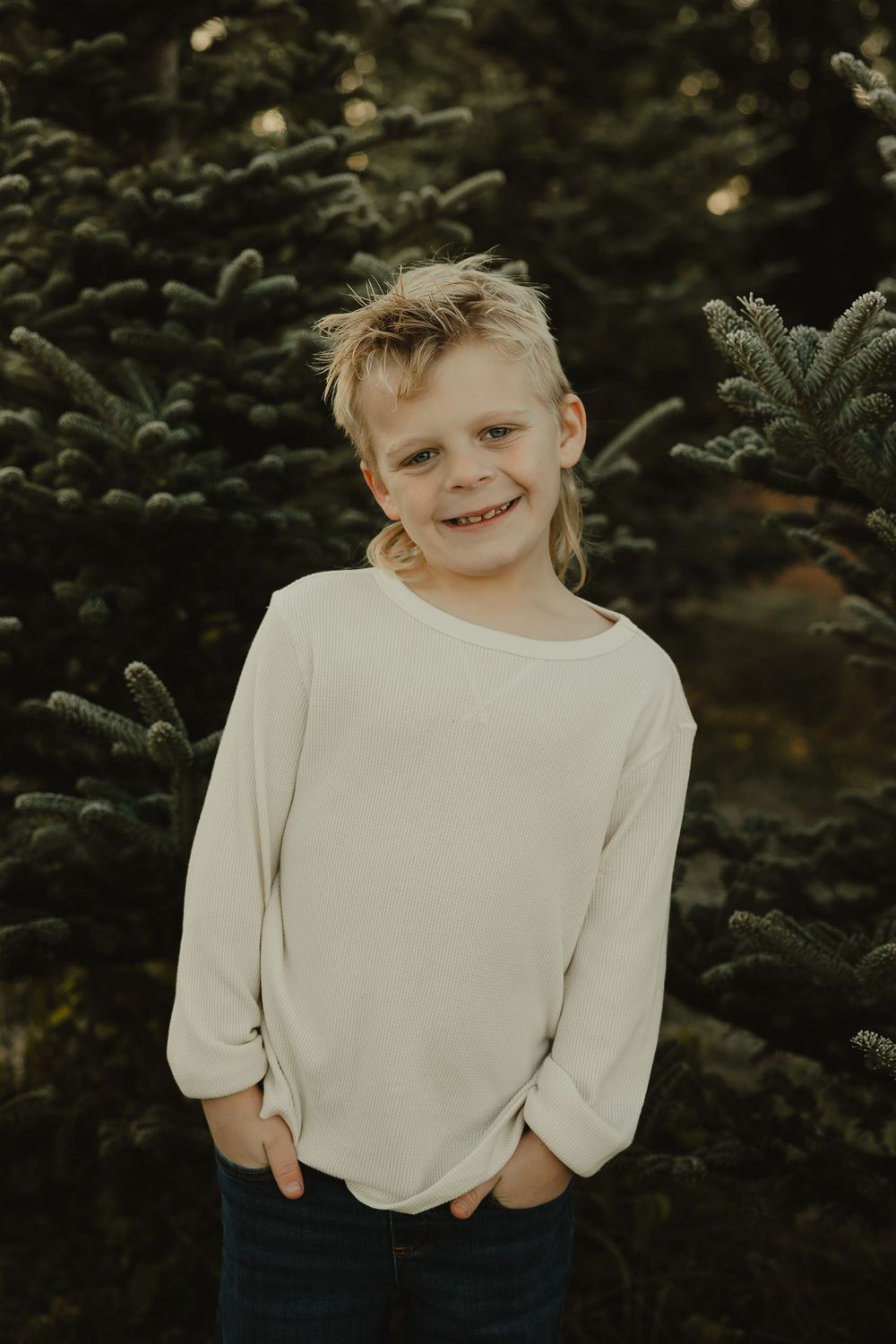 Child in cream sweater smiling warmly against evergreen tree backdrop.