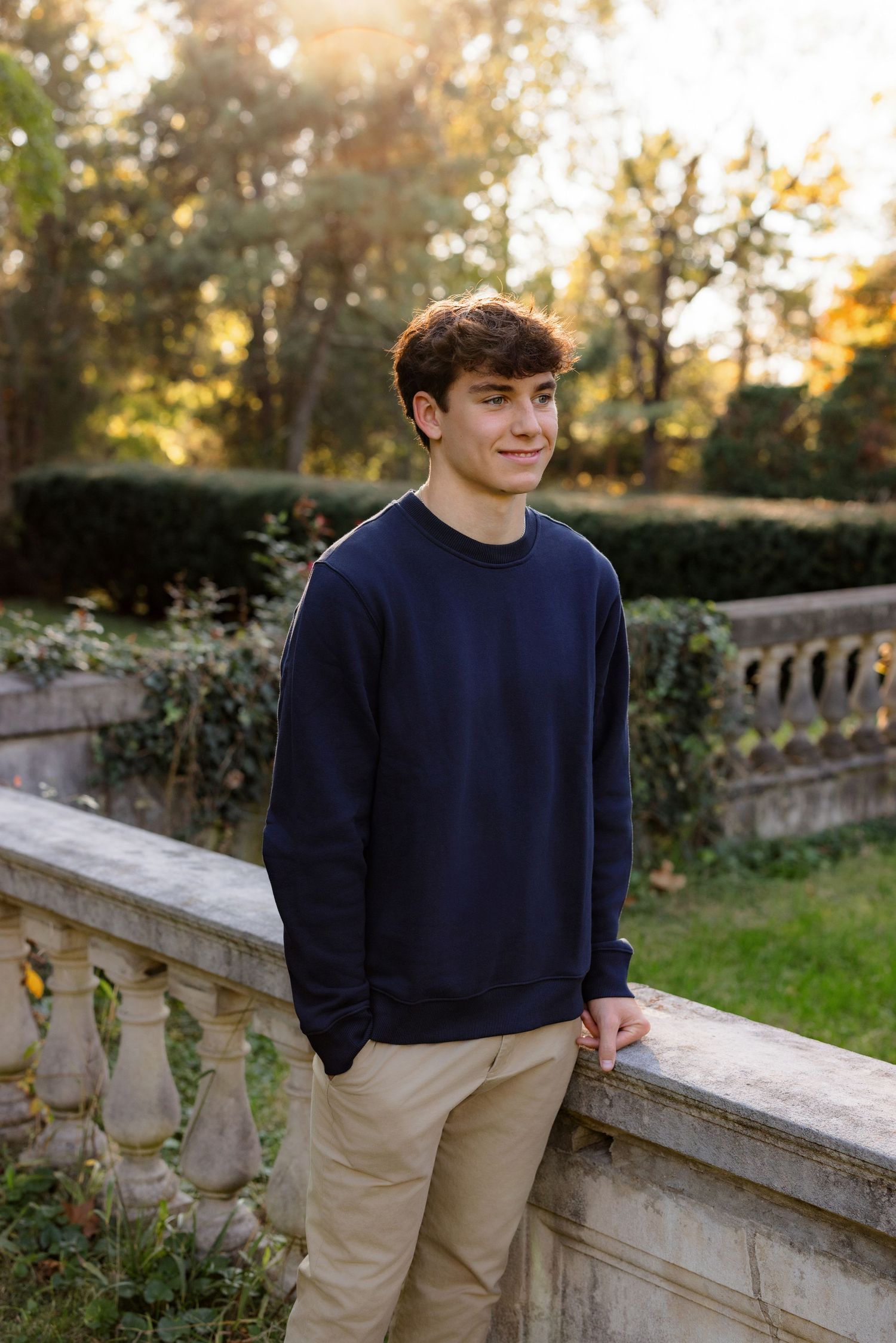 A person in a navy sweatshirt and khaki pants poses along a stone balustrade in a garden at sunset.
