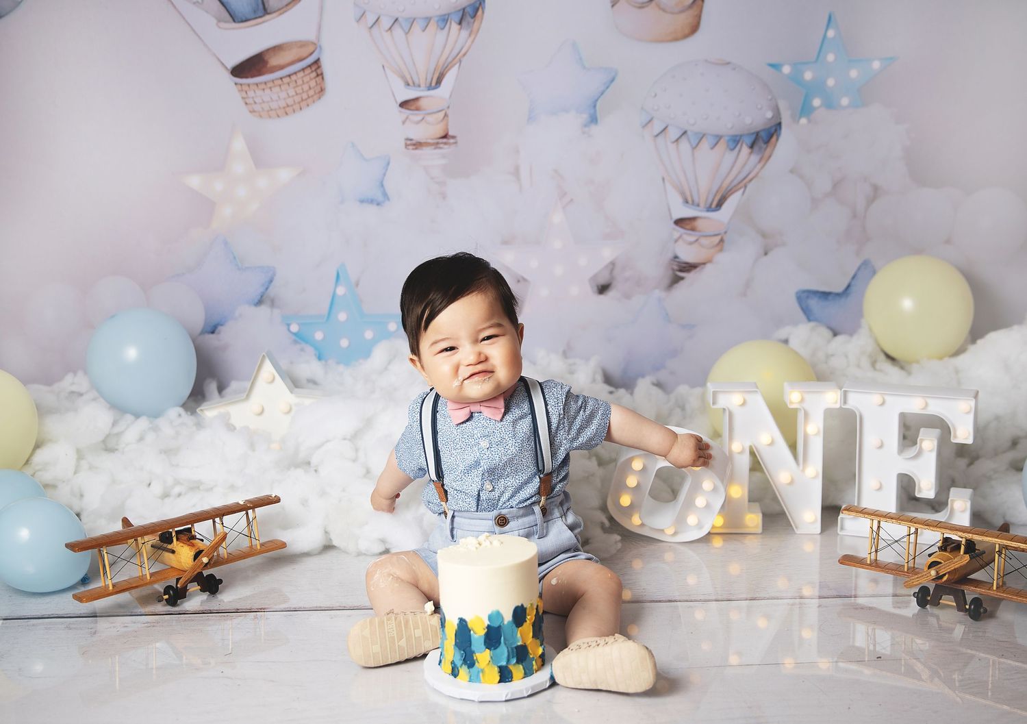 Baby in blue suspenders sits with birthday cake amid vintage planes, clouds, stars and pastel balloon decorations.