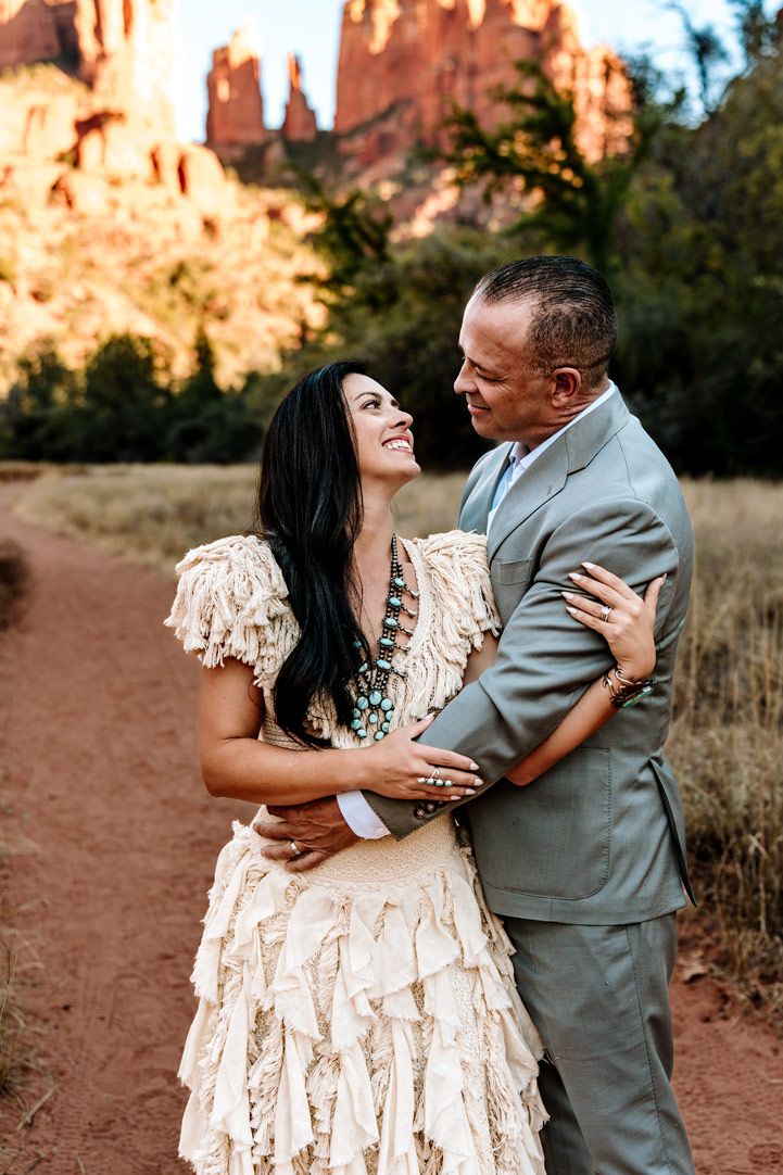 A loving couple embraces on a Sedona red dirt trail with majestic sandstone formations glowing in the desert sunset.