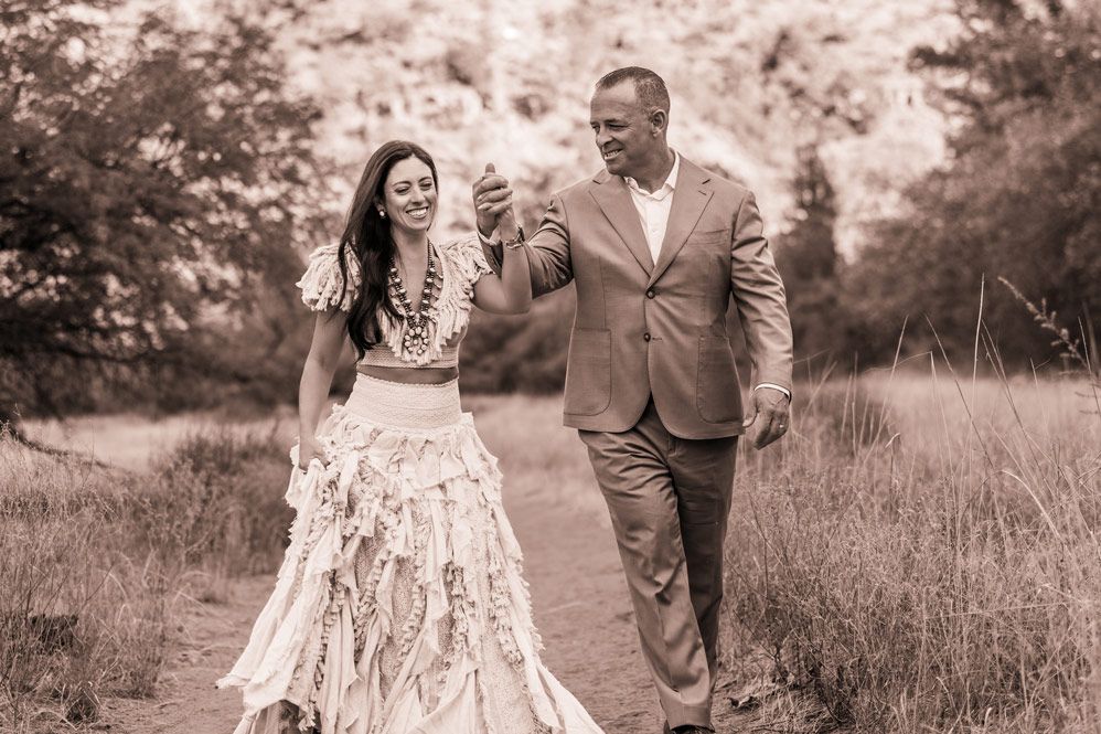 A joyful couple in formal attire share a laugh while walking through a sunlit field.