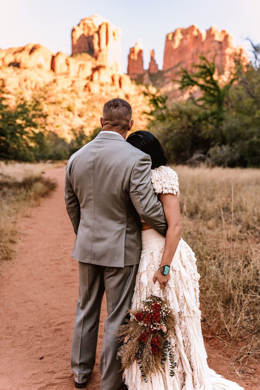A couple in formal attire stands on a Sedona red dirt path admiring towering desert rock formations at sunset.