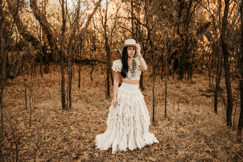 A woman in a flowing white lace dress stands in a golden autumn forest.